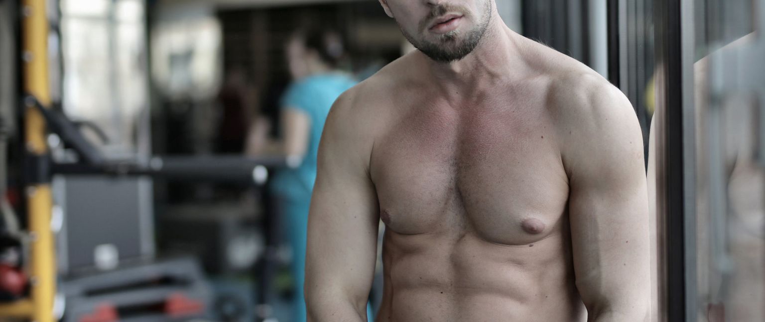 Close-up of a muscular, shirtless man's torso and chest in a gym.