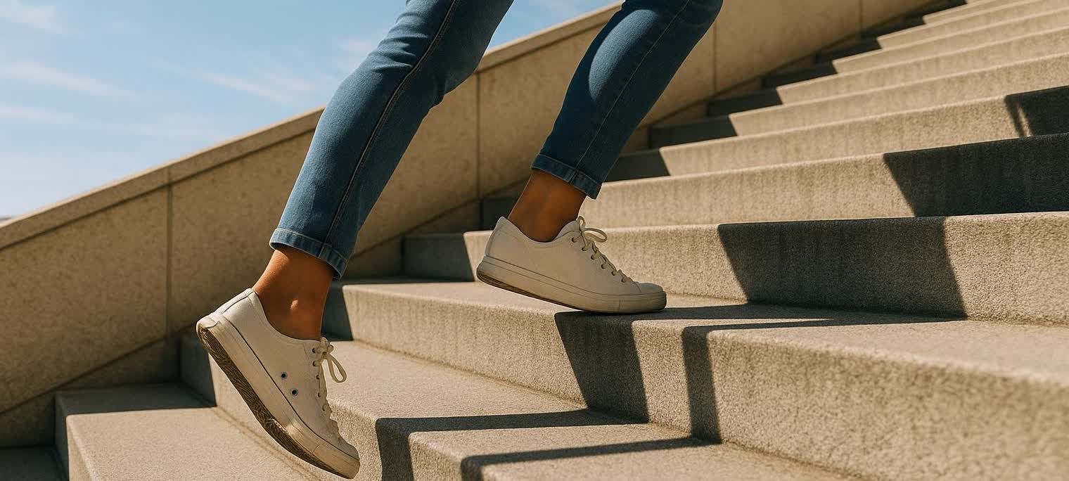 A low-angle view of a person's legs, clad in blue jeans and white sneakers, confidently walking up a set of wide, tan outdoor stairs with strong sunlight casting sharp shadows.