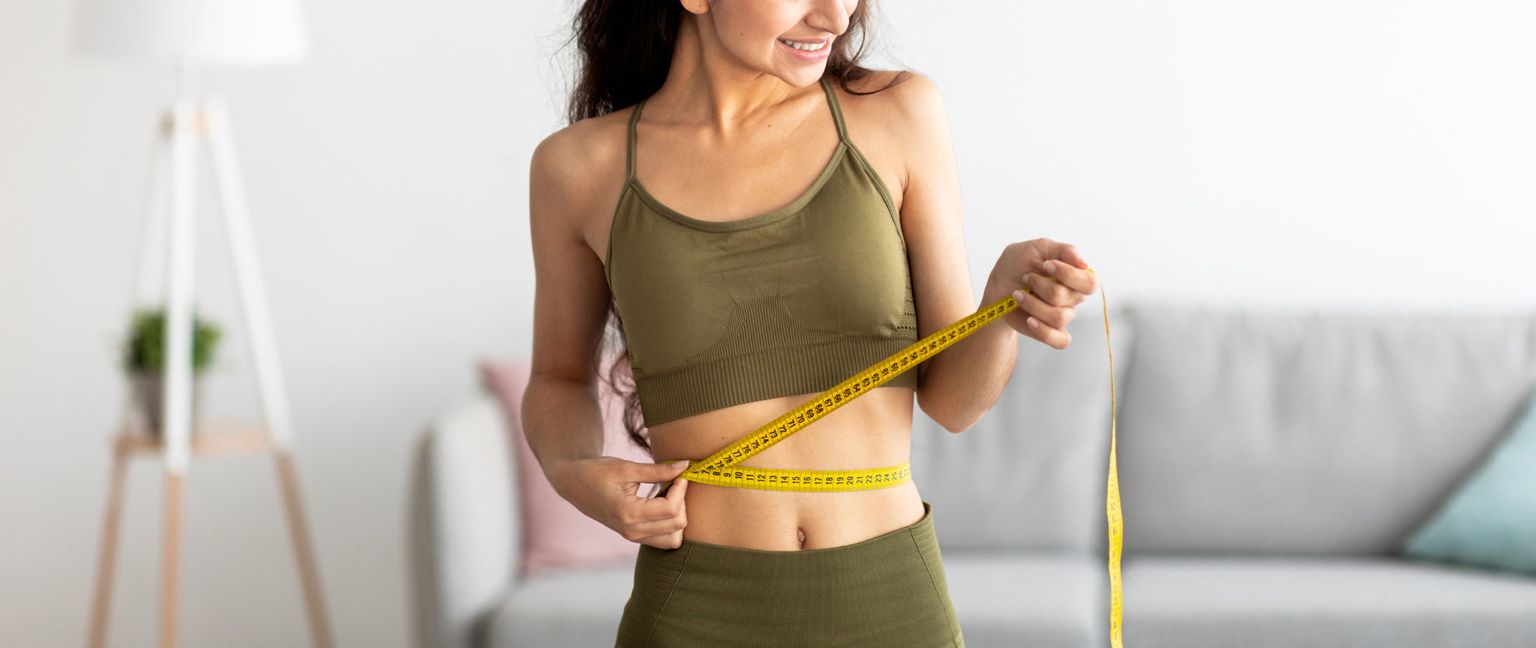 Close up image of a woman in activewear measuring their waist with a yellow measuring tape.