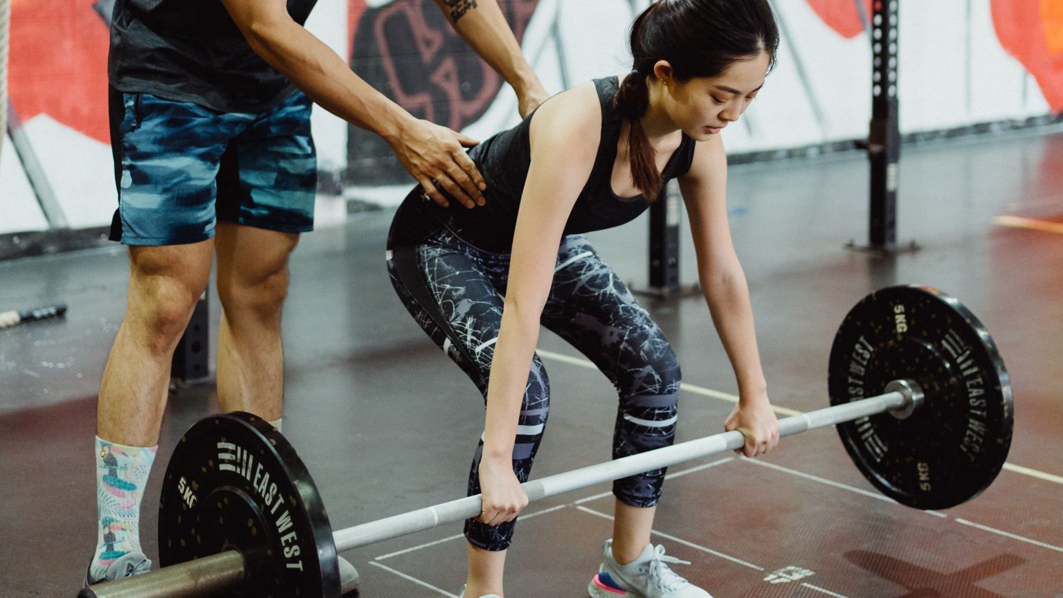 A woman in a black tank top and patterned leggings prepares to deadlift a barbell while a coach with his hand on her lower back instructs her.