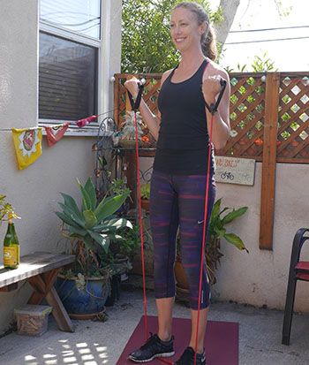 A woman smiles while exercising with red resistance bands outdoors on a sunny day.