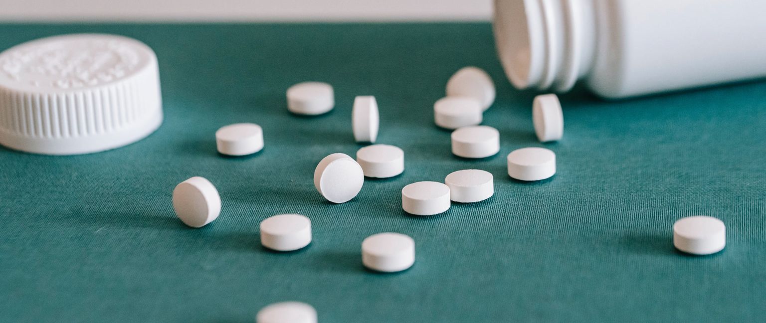 A white pill bottle is tipped on its side, and round white pills spill out onto a teal-colored surface.