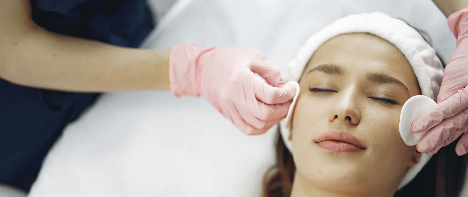 A woman with her eyes closed receives a facial treatment at a spa. A hand in a pink glove gently cleans her face with a cotton pad.