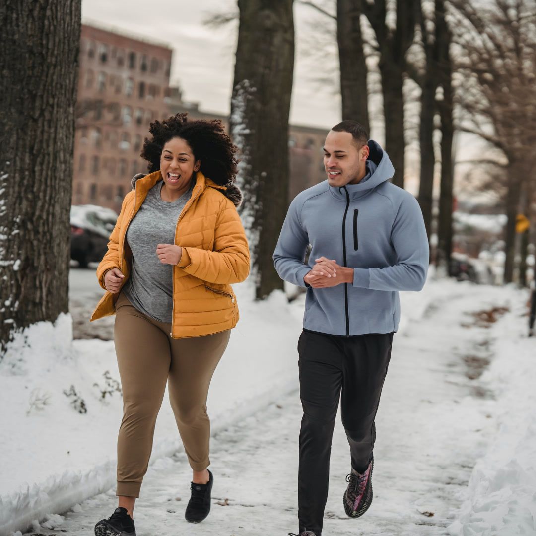Two people joyfully running on a snow-covered sidewalk lined with trees and buildings.