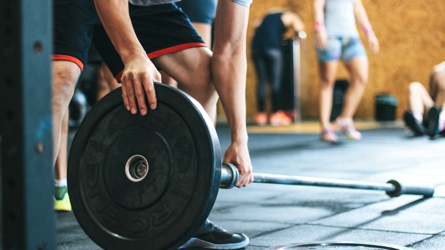 A man in athletic wear stands at a squat rack, his hands placing a large black weight plate onto the end of a silver barbell.
