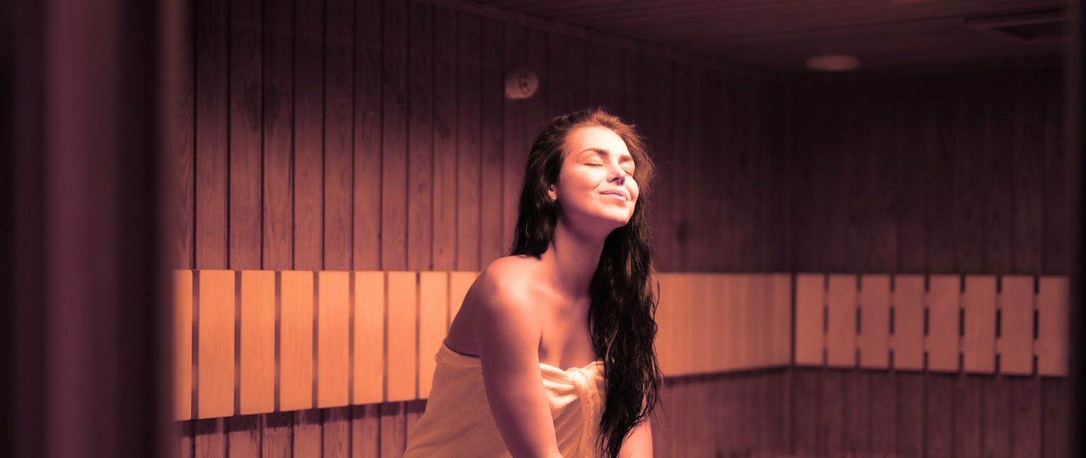 A young woman with long dark hair and closed eyes is smiling happily while relaxing in a sauna, wrapped in a light-colored towel.