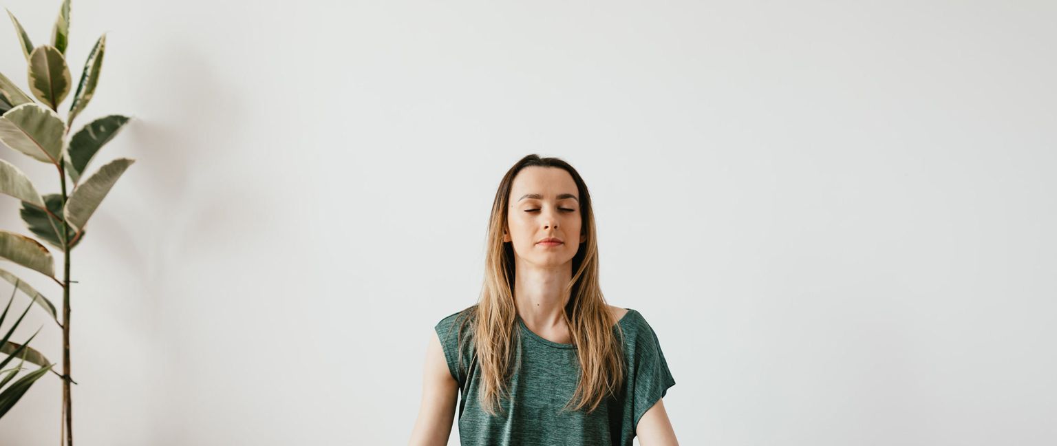 A woman with her eyes closed meditates in a room with a white wall and a plant.