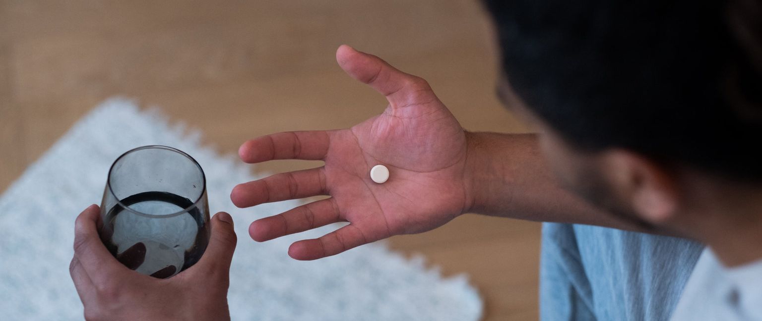 A white pill rests in a man's outstretched palm, while his other hand holds a glass of water.