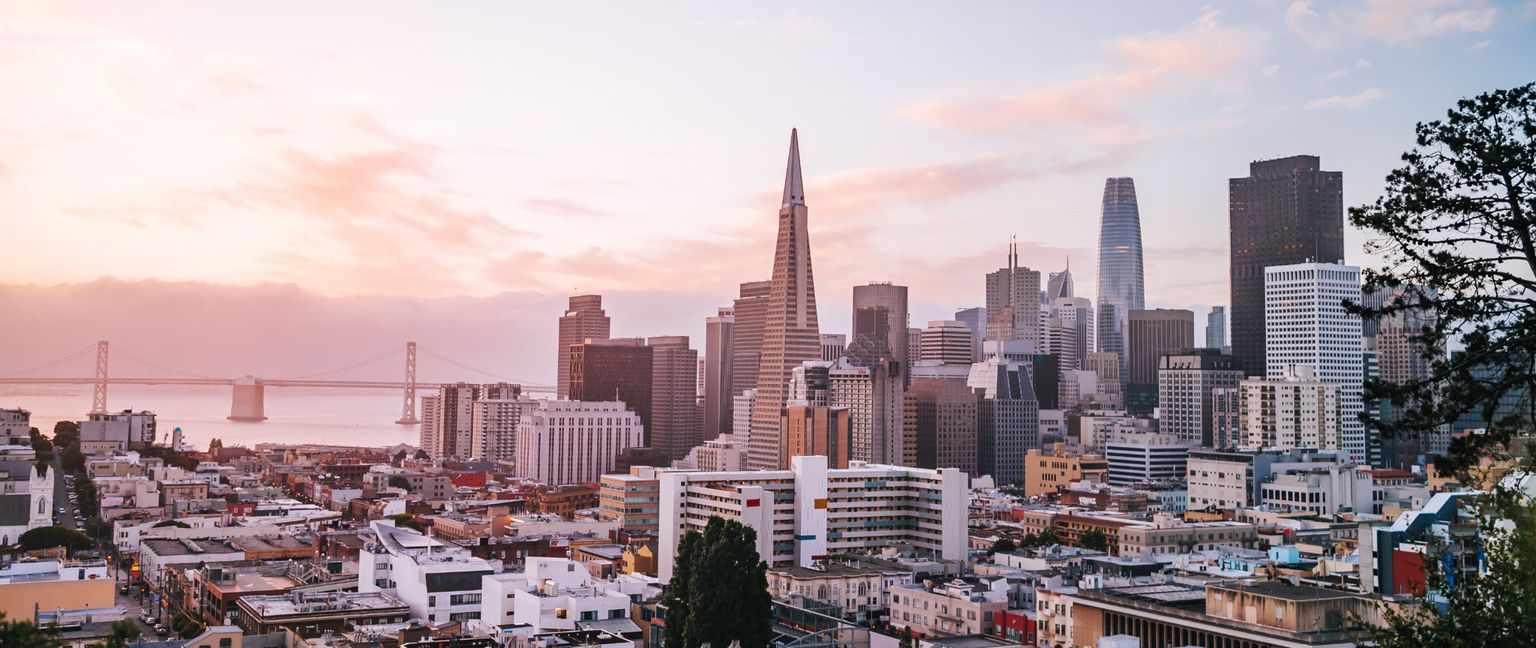 A panoramic view of the San Francisco skyline and the Bay Bridge at sunset, with the Transamerica Pyramid and Salesforce Towe visible among other buildings, with residential buildings in the foreground.