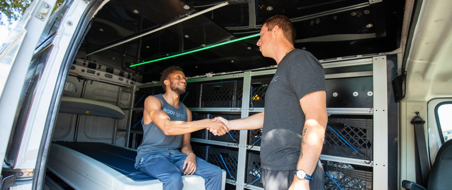 Two men shake hands inside the back of a customized van.