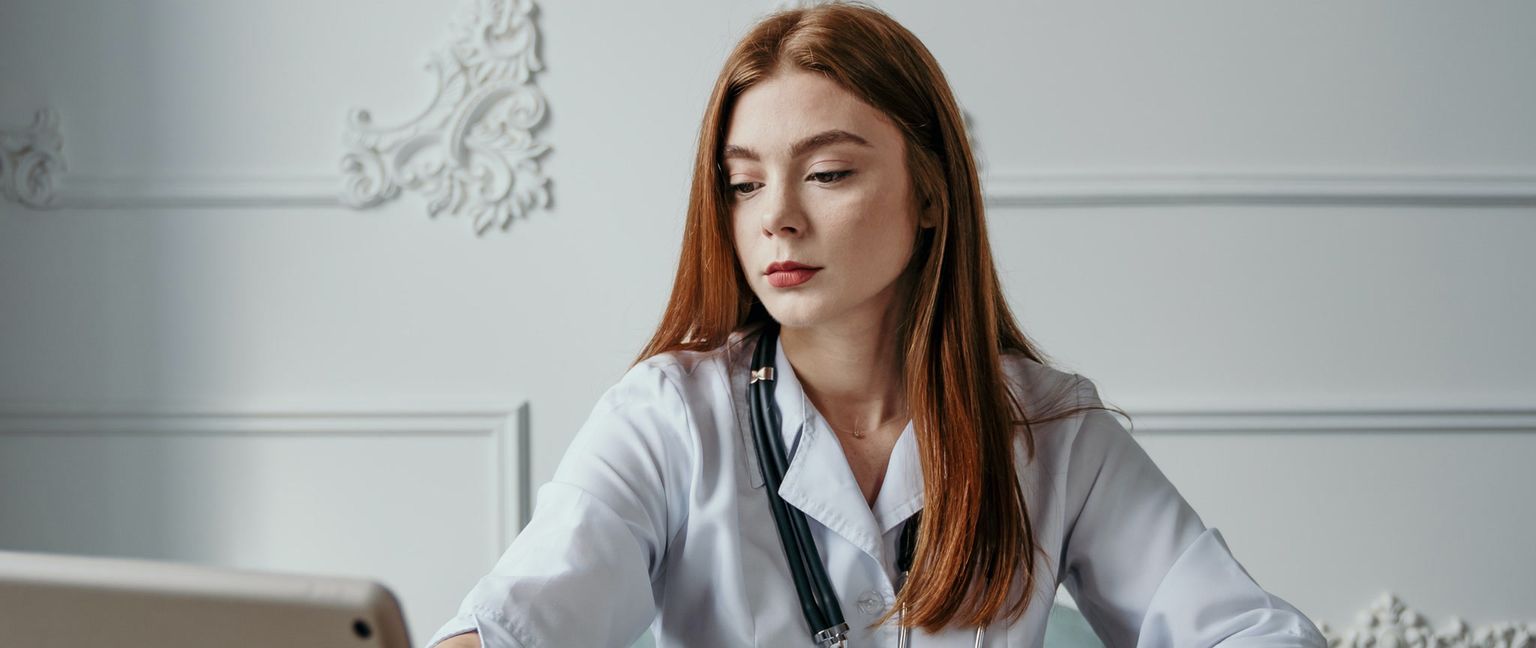 A seated female doctor with long red hair and a stethoscope around her neck looks down at a tablet she is using, against a white background with molding.