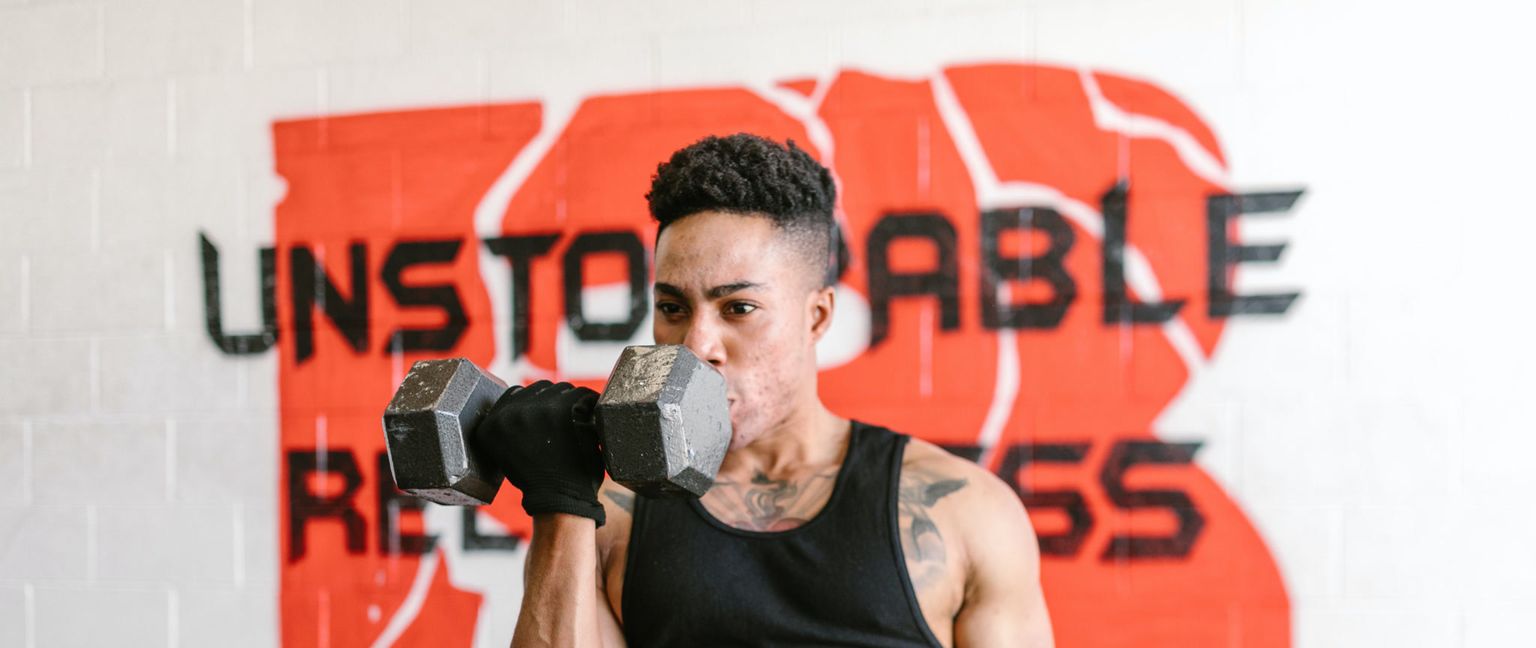 A determined young man with short, dark hair and tattoos wears a black tank top and a black glove as he lifts a heavy dumbbell.