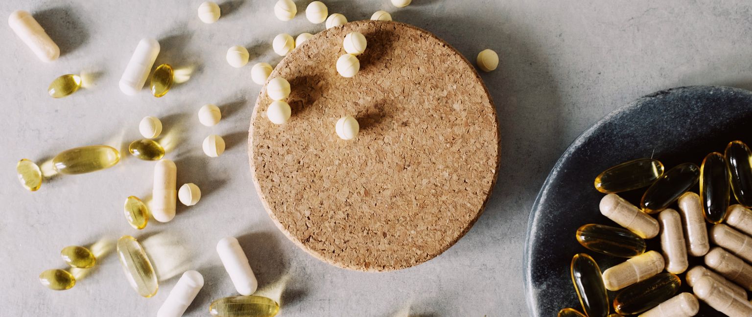 An overhead view of various vitamins and supplements scattered on a gray surface, including white capsules, yellow softgels, small round tablets, and a dark bowl filled with supplements.