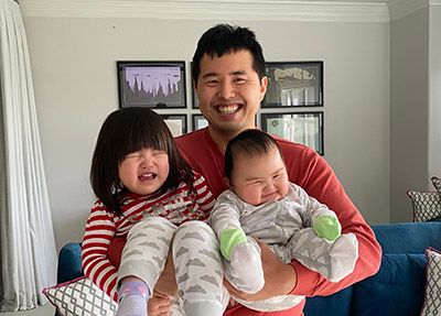 A father smiles broadly while holding his crying toddler daughter on his left and his smiling infant son on his right. The father is wearing a red shirt, the toddler has short dark hair and is wearing a red and white striped shirt and cloud-patterned leggings, and the baby is wearing a grey and white cloud-patterned onesie.