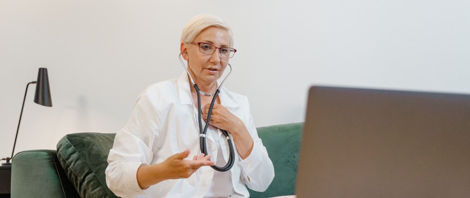 A female doctor in a white coat sits on a Couch with a stethoscope around her neck talking to a patient on a computer.