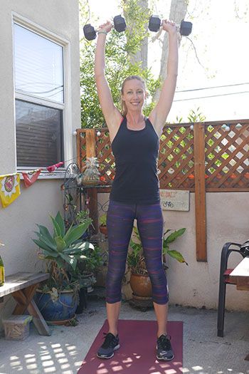 A woman performs an overhead dumbbell press on a mat outdoors on a sunny day.