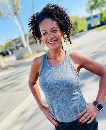 A woman with curly hair smiles at the camera, wearing a grey tank top and purple smartwatch, with her hands on her hips.