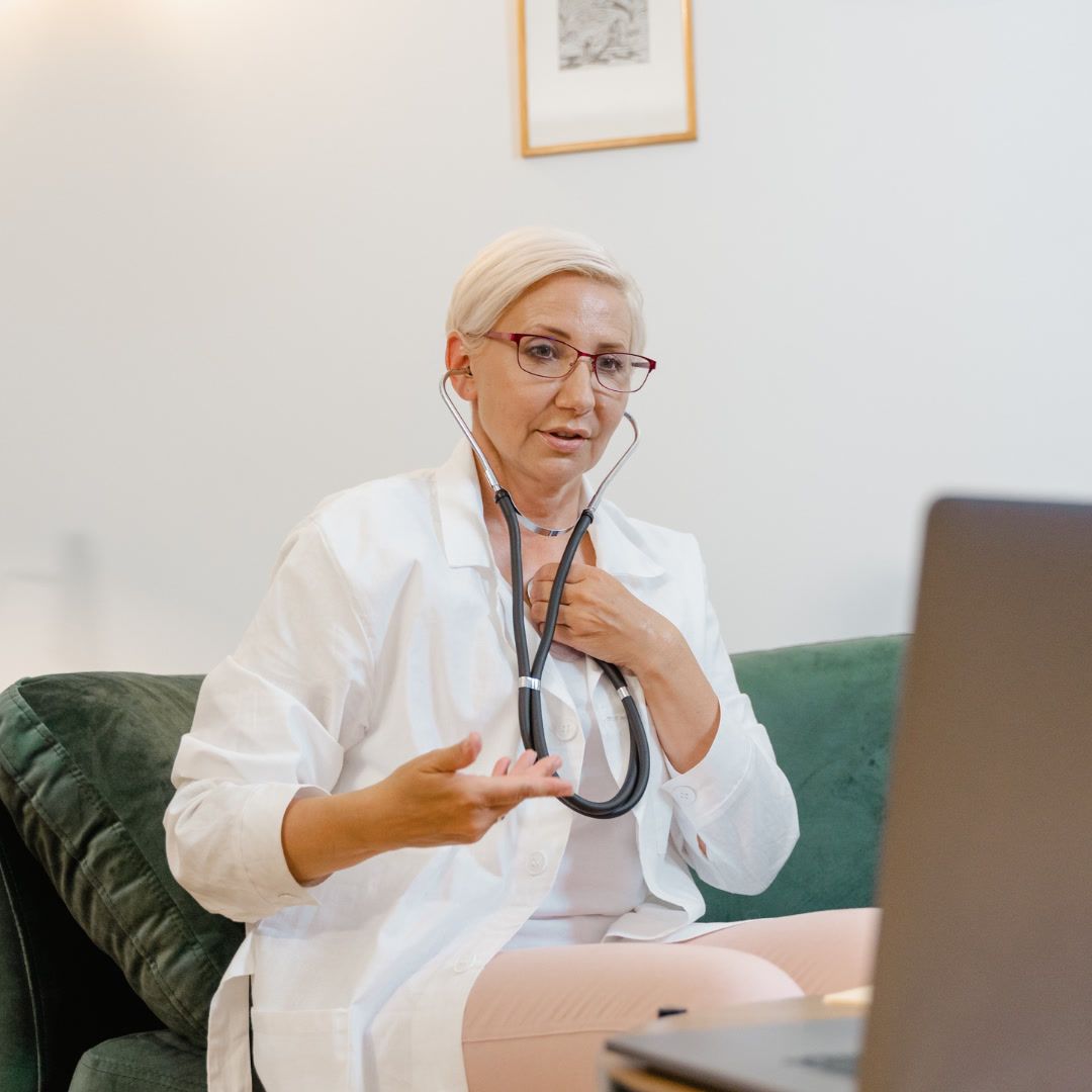A female doctor wearing a white coat and stethoscope sitting on a couch and on a video call with a patient on her laptop.