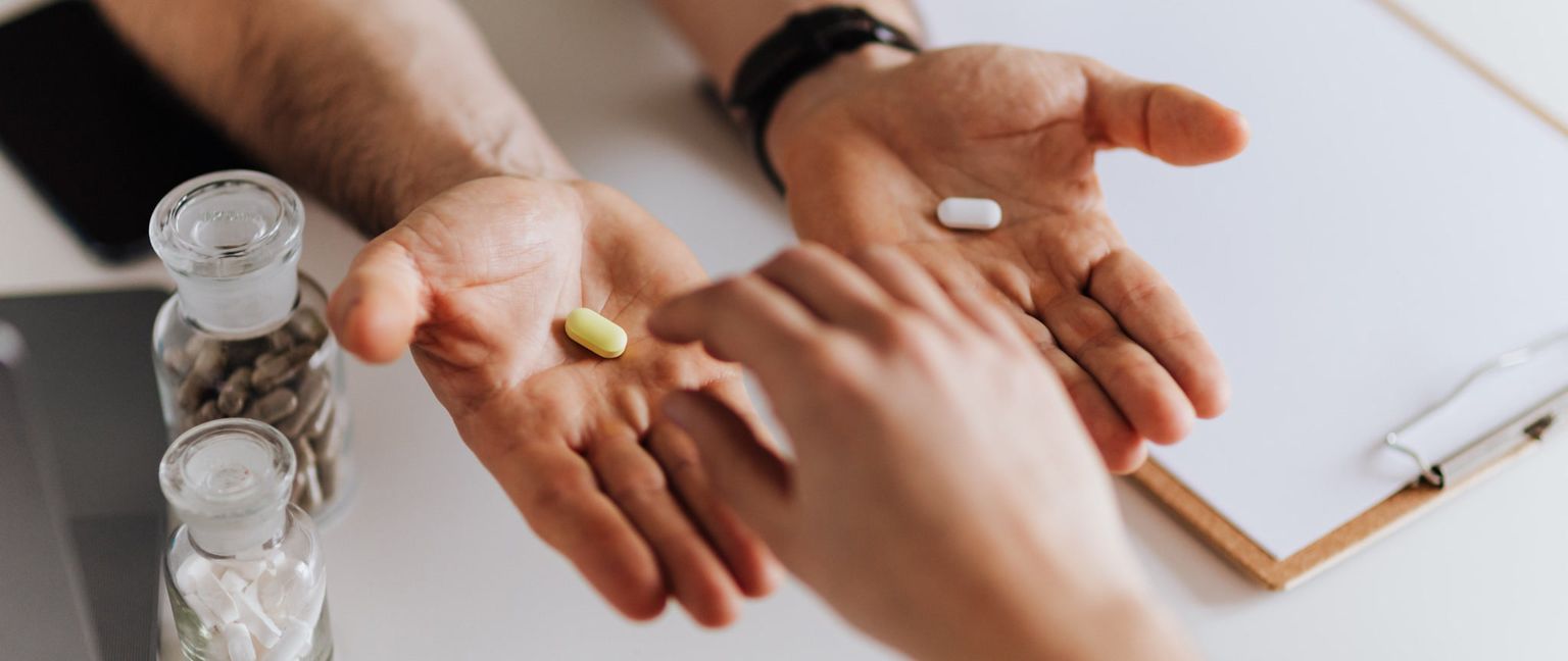 Close-up of two hands holding a yellow pill and a white pill, suggesting a choice between two options.