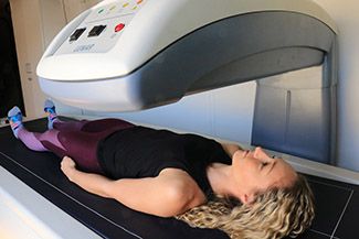 A woman lies still on a table under a large white machine, likely for a bone density scan.