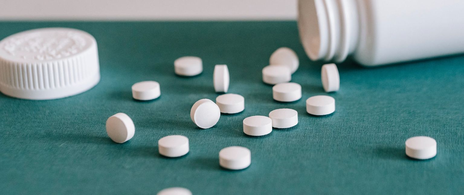 Several white pills are scattered on a teal background, with a white bottle cap and a white bottle visible in the background.