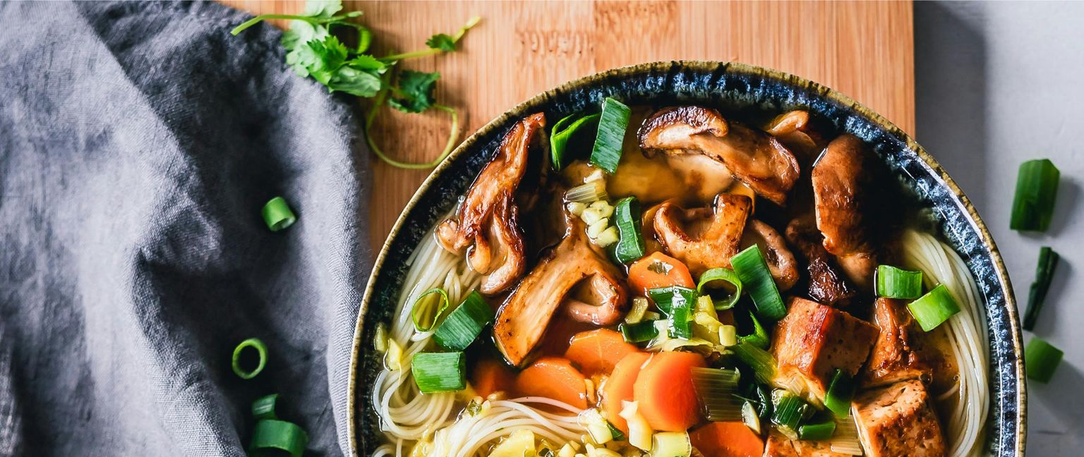 Close-up view of a bowl of vegetarian noodle soup with mushrooms, carrots, tofu, and green onions.