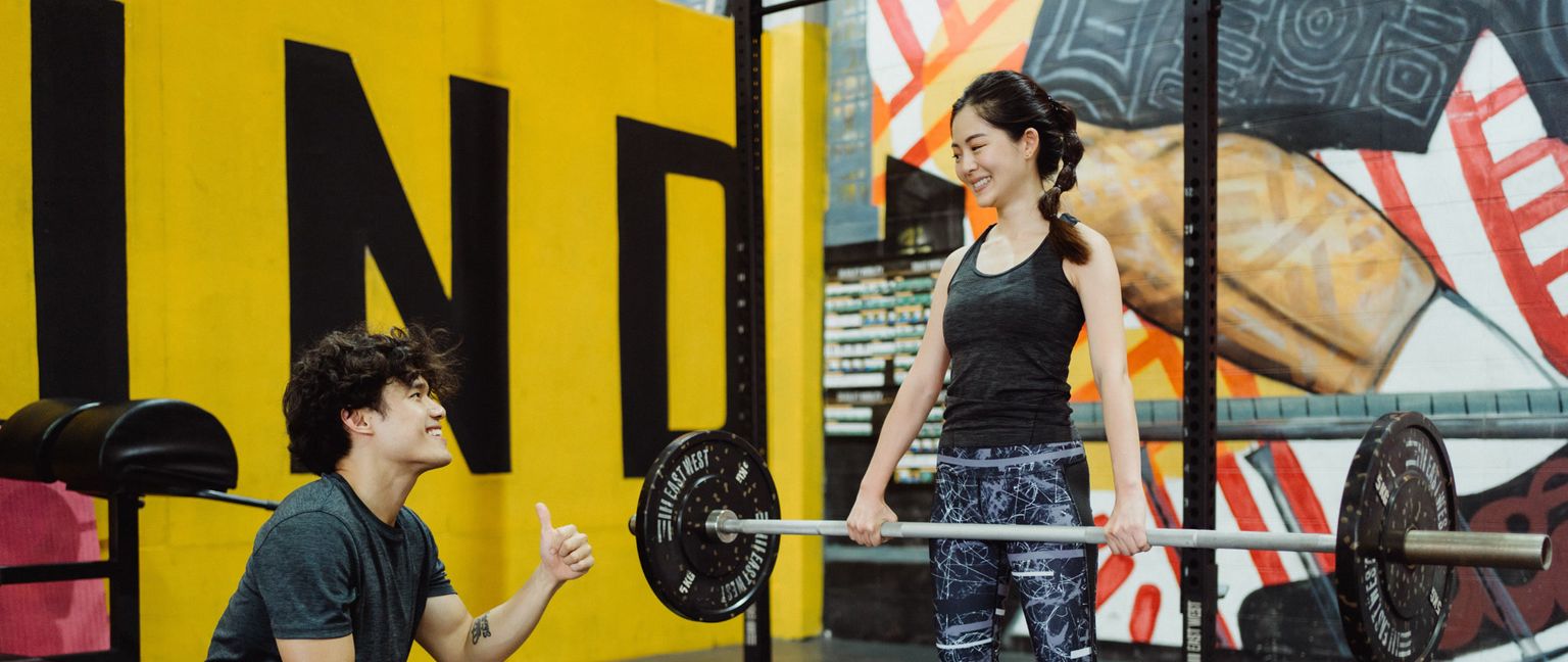 A man gives a woman a thumbs up while she holds a barbell in a brightly painted gym.