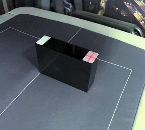 A black Block held down with white plastic, with a red crosshair on a white square on the top. Calibration lines are visible underneath it on a grey surface.