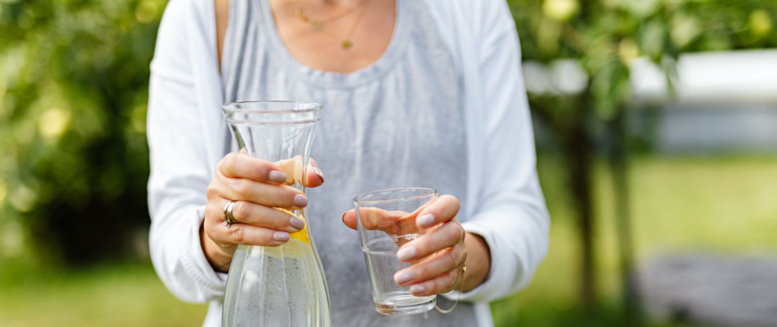 A woman holding a glass carafe filled with water and lemon slices in one hand and an empty glass in the other. She is wearing a white cardigan and a grey shirt. A green outdoor background is blurred behind her.
