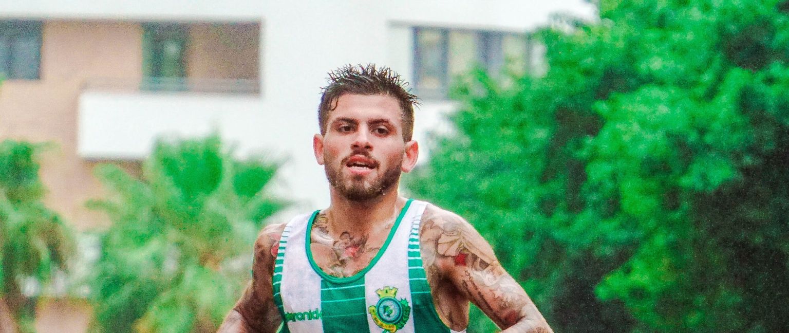 A man in a green and white running singlet runs towards the camera with trees and buildings in the background.