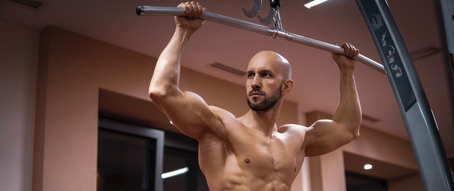 A muscular man with a shaved head and beard is pulling down on a cable machine attachment, working his back and biceps.